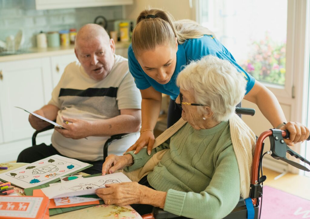 A compassionate caregiver leaning over to talk warmly with an elderly woman in a wheelchair during an art activity, with a senior man sitting in the background.