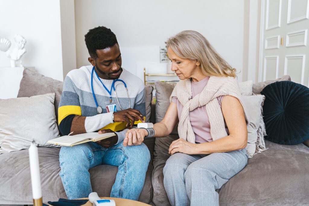 A male nurse with a stethoscope checking a senior woman's vital signs using a wrist blood pressure monitor on a living room sofa.