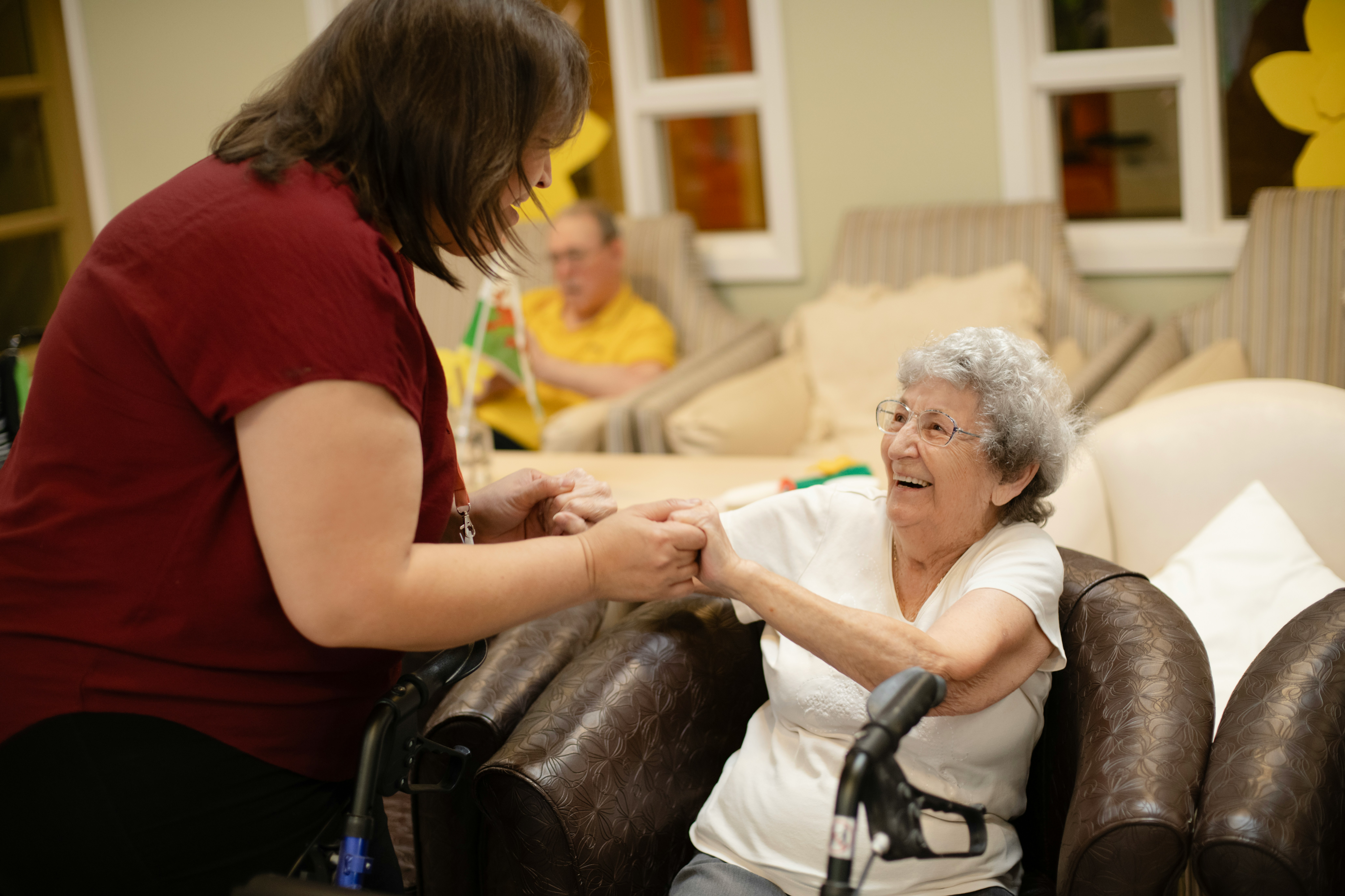 A female caregiver holding hands and sharing a warm, joyful conversation with a smiling senior woman seated in an armchair.
